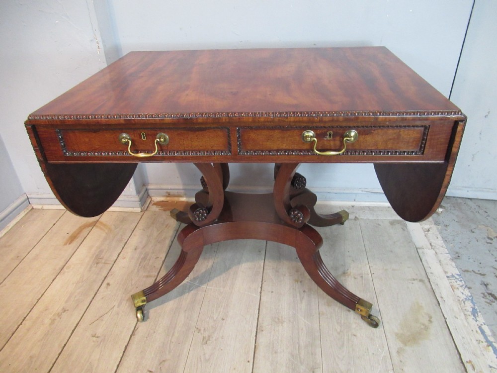 An early 19th-century mahogany sofa table featuring two drop leaf flaps, two front drawers with brass handles, twin scroll supports, and reeded legs with brass castors.