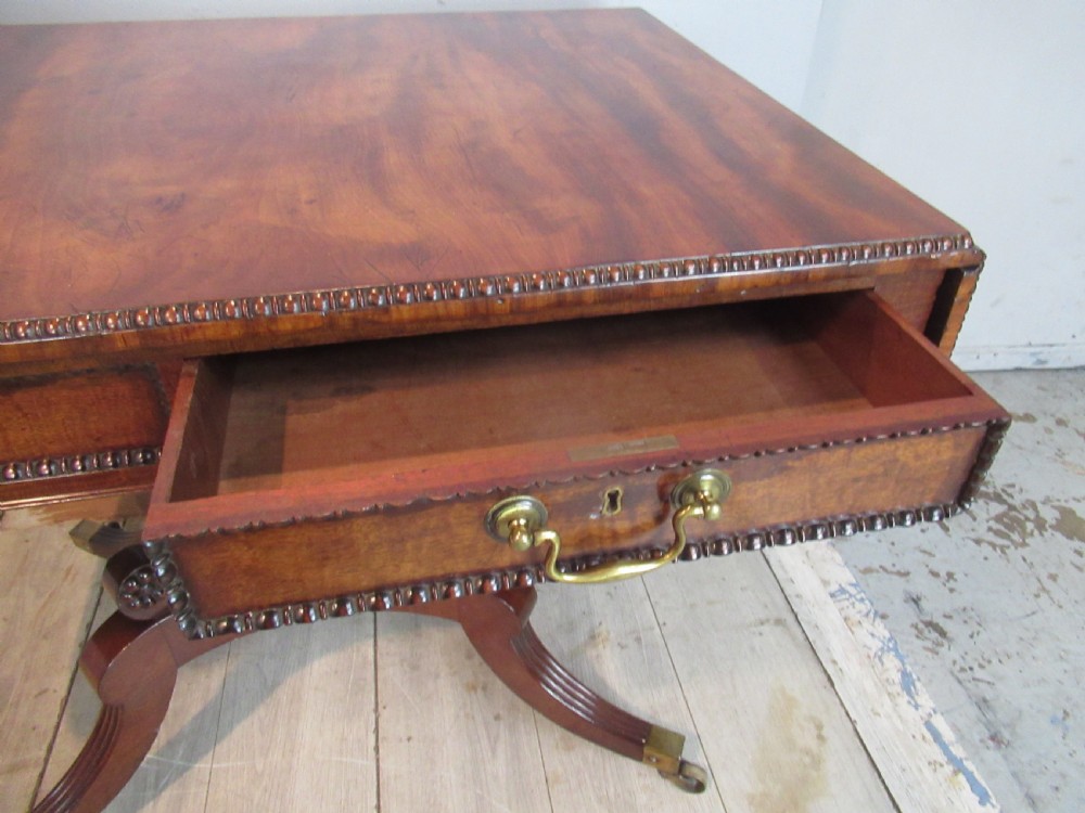 An early 19th-century mahogany sofa table featuring two drop leaf flaps, two front drawers with brass handles, twin scroll supports, and reeded legs with brass castors. View IX