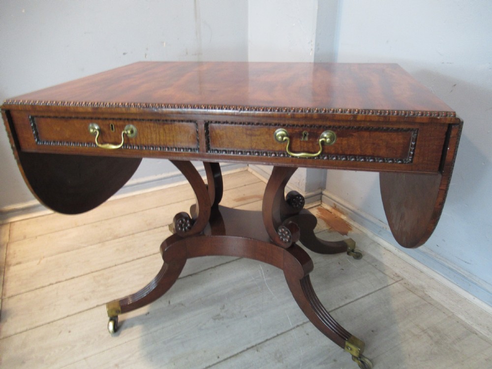 An early 19th-century mahogany sofa table featuring two drop leaf flaps, two front drawers with brass handles, twin scroll supports, and reeded legs with brass castors. View XI