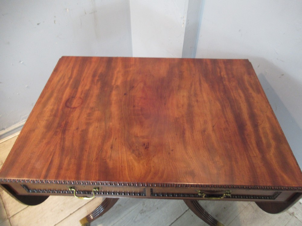 An early 19th-century mahogany sofa table featuring two drop leaf flaps, two front drawers with brass handles, twin scroll supports, and reeded legs with brass castors. View XII