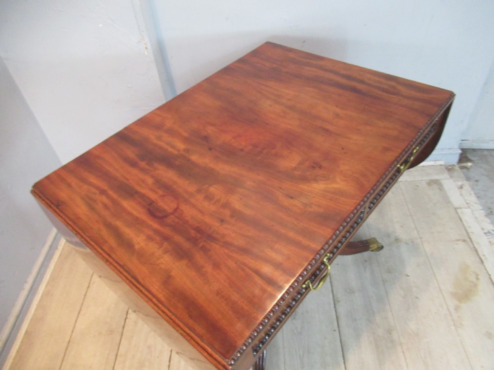 An early 19th-century mahogany sofa table featuring two drop leaf flaps, two front drawers with brass handles, twin scroll supports, and reeded legs with brass castors. View XIII