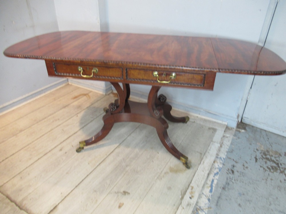 An early 19th-century mahogany sofa table featuring two drop leaf flaps, two front drawers with brass handles, twin scroll supports, and reeded legs with brass castors. View XV
