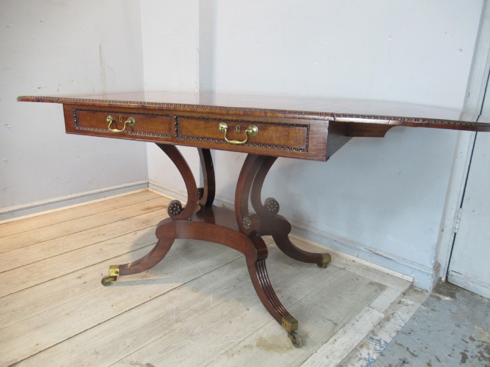 An early 19th-century mahogany sofa table featuring two drop leaf flaps, two front drawers with brass handles, twin scroll supports, and reeded legs with brass castors. View XVI