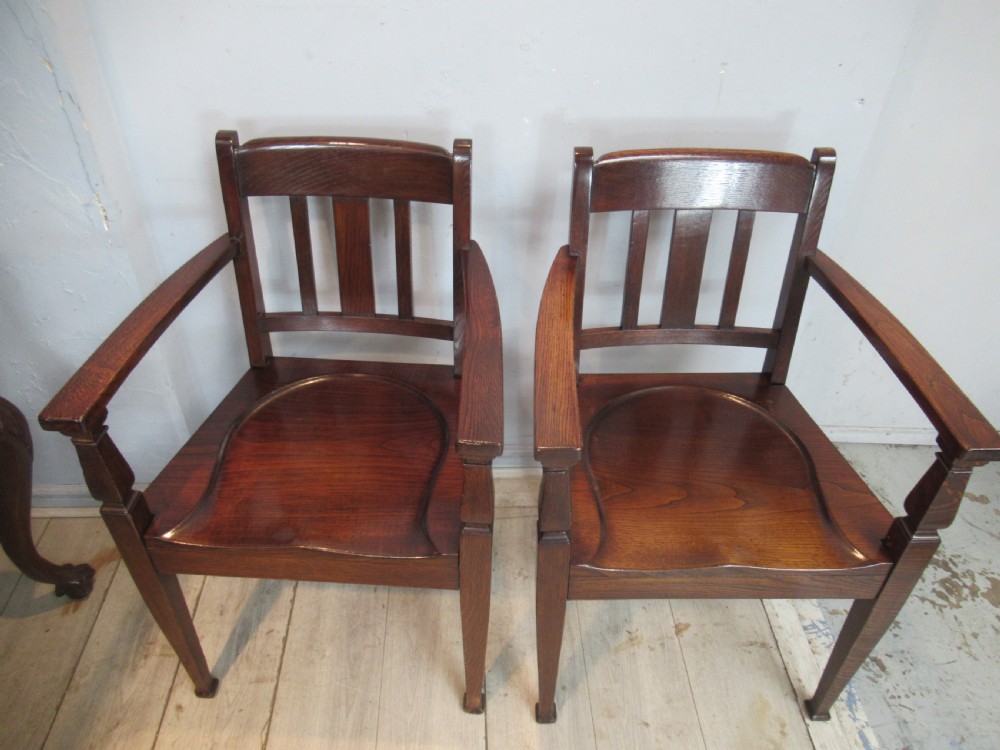 A pair of dark oak Arts and Crafts armchairs featuring vertical back slats, contoured solid timber seats, and straight tapered legs.