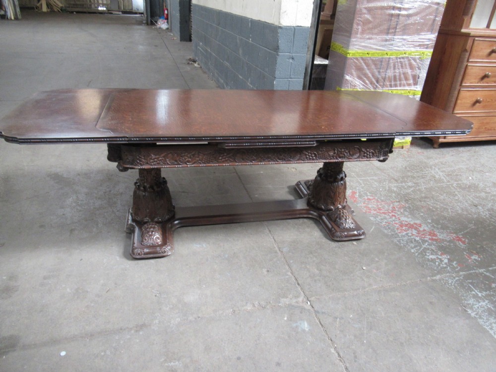 A dark carved oak extending dining table featuring ornate pedestal supports and a carved apron.