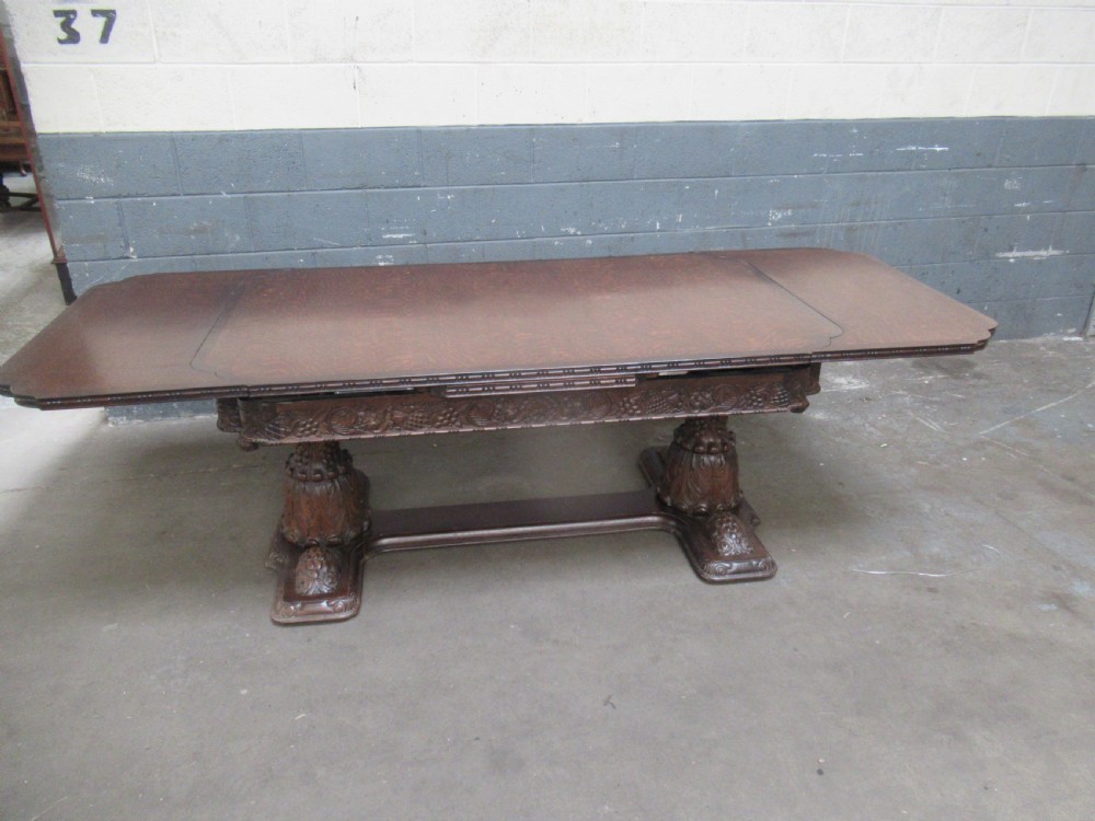 A dark carved oak extending dining table featuring ornate pedestal supports and a carved apron. View II