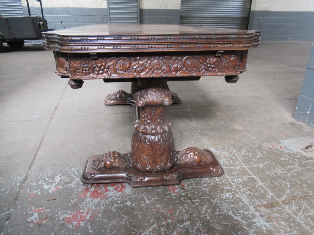 A dark carved oak extending dining table featuring ornate pedestal supports and a carved apron. View XI