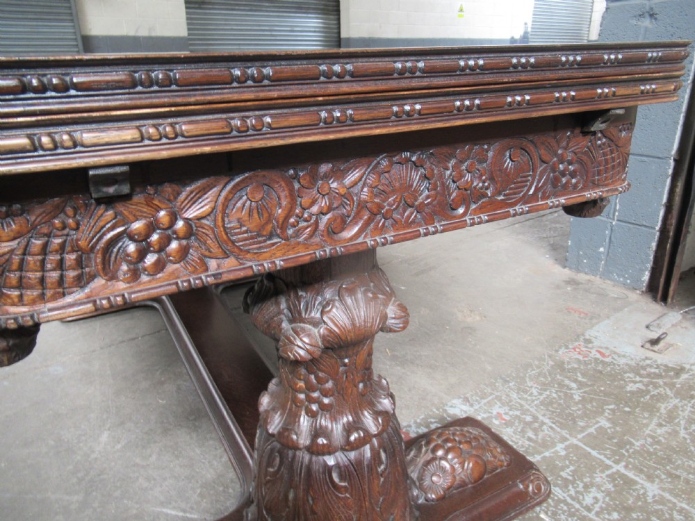 A dark carved oak extending dining table featuring ornate pedestal supports and a carved apron. View XV