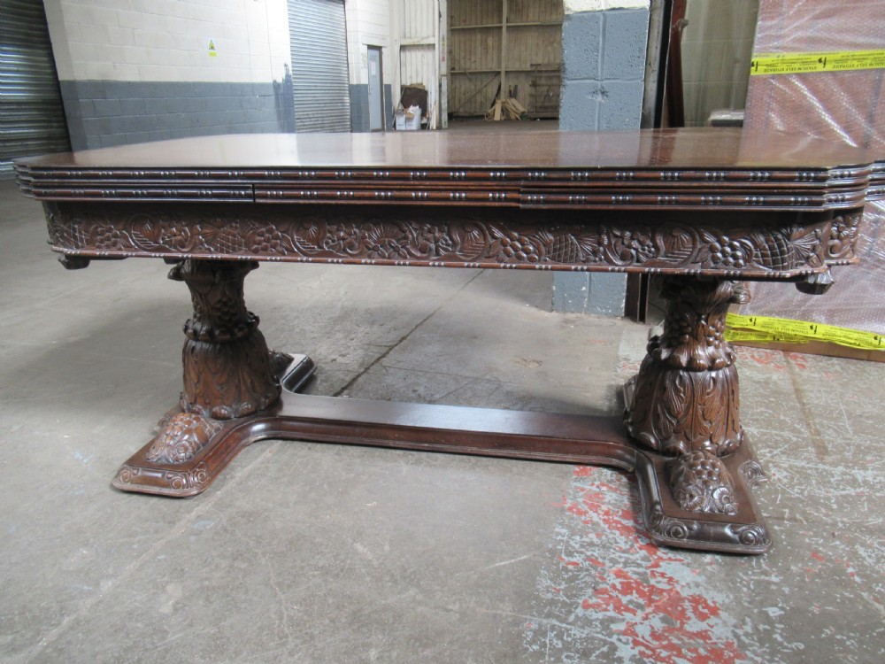A dark carved oak extending dining table featuring ornate pedestal supports and a carved apron. View XVI