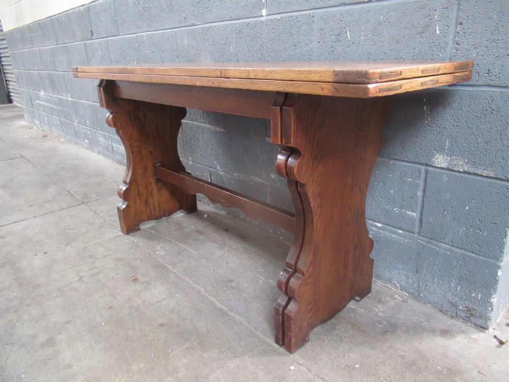 A rectangular 1920s oak fold-over dining table featuring panelled timber top sections, brass hinges, and shaped trestle-style slab supports. View VIII