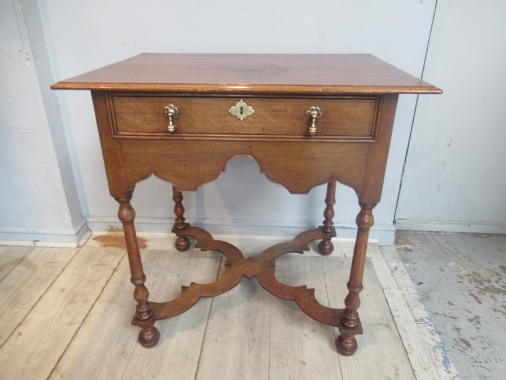 An antique warm brown oak side table featuring a single drawer with brass drop handles, a shaped apron, turned legs, and an X-frame stretcher.