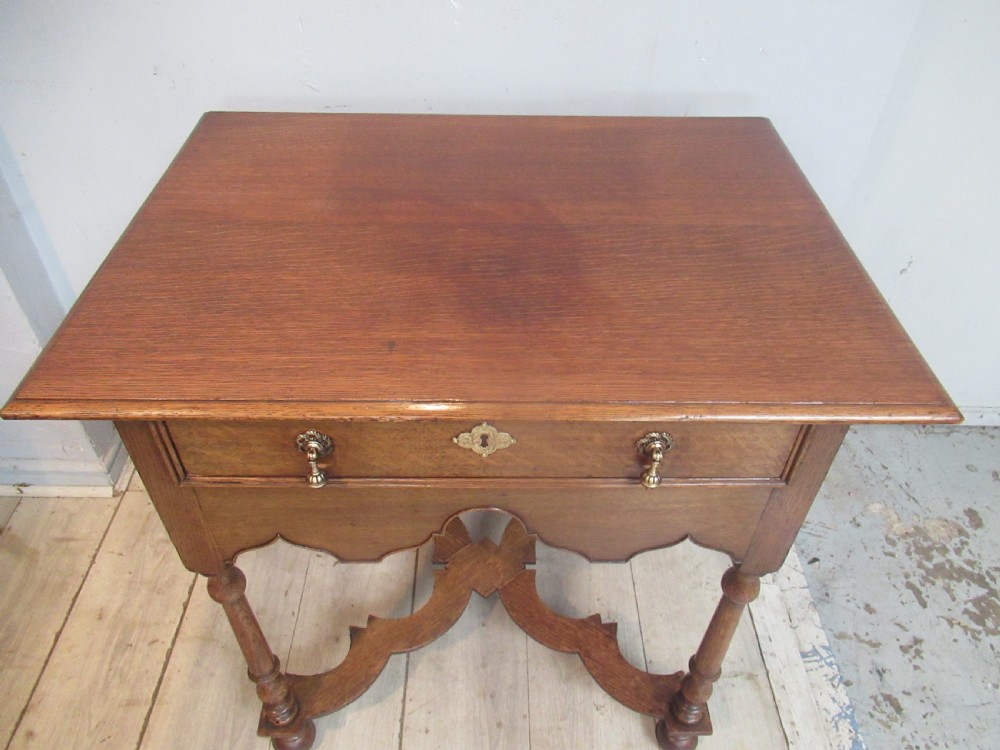 An antique warm brown oak side table featuring a single drawer with brass drop handles, a shaped apron, turned legs, and an X-frame stretcher. View III