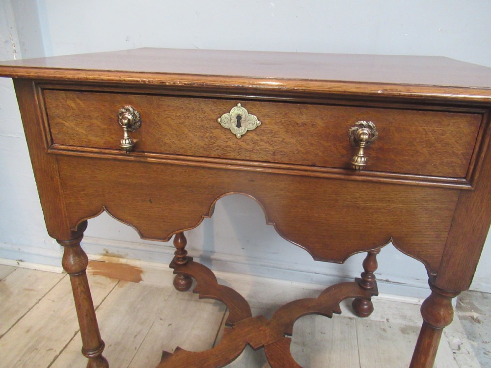 An antique warm brown oak side table featuring a single drawer with brass drop handles, a shaped apron, turned legs, and an X-frame stretcher. View V