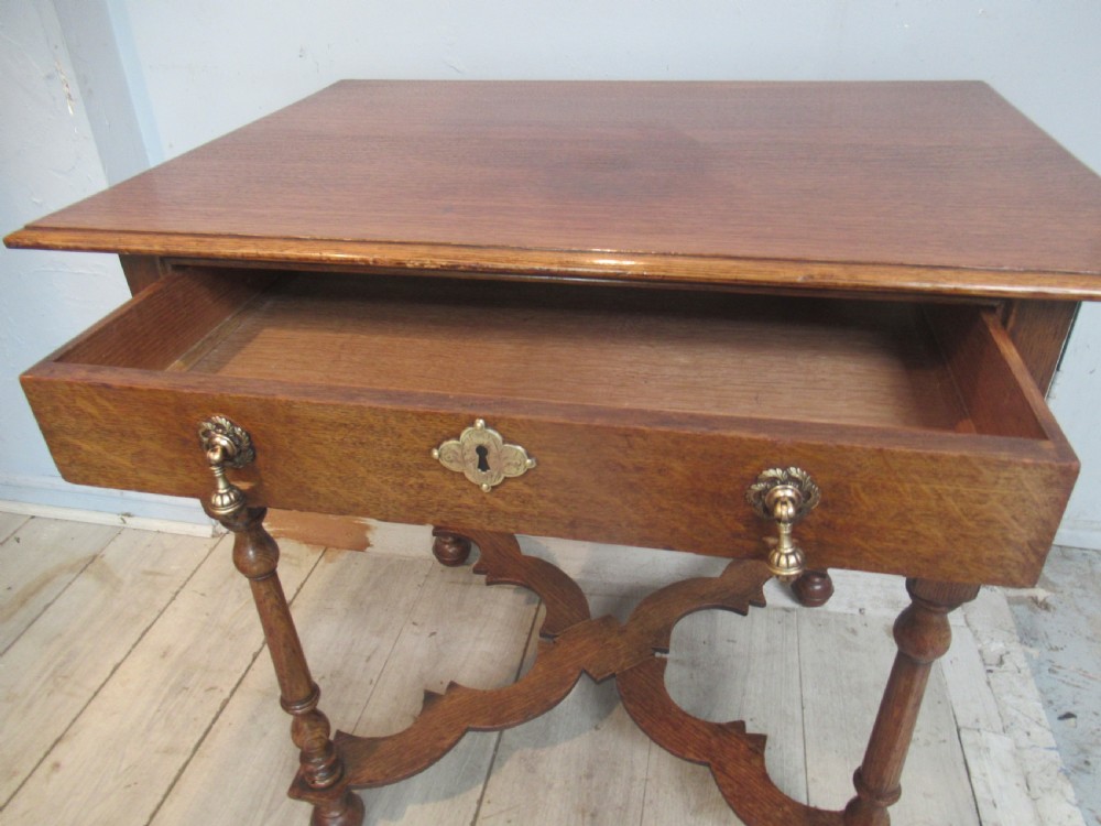 An antique warm brown oak side table featuring a single drawer with brass drop handles, a shaped apron, turned legs, and an X-frame stretcher. View VI