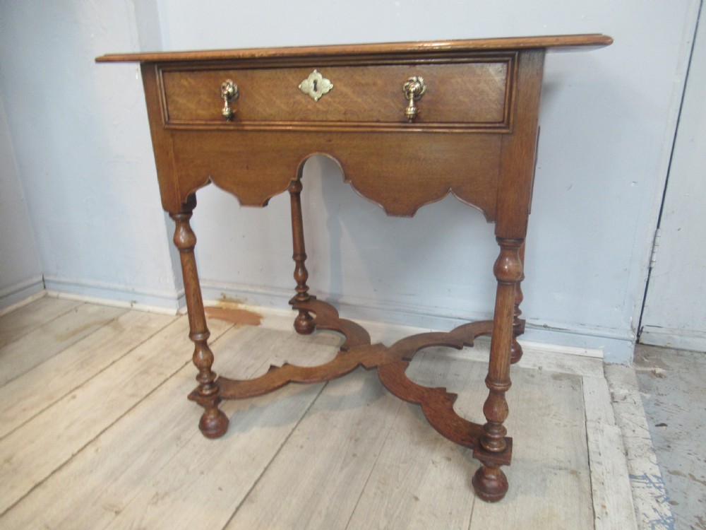 An antique warm brown oak side table featuring a single drawer with brass drop handles, a shaped apron, turned legs, and an X-frame stretcher. View X