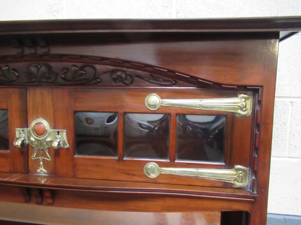 An Arts and Crafts period mahogany sideboard featuring leaded glass doors, brass strap hinges, a central carved tree motif, and a mirror back. View V