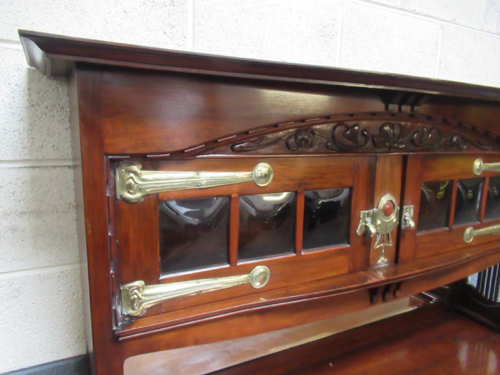 An Arts and Crafts period mahogany sideboard featuring leaded glass doors, brass strap hinges, a central carved tree motif, and a mirror back. View VII