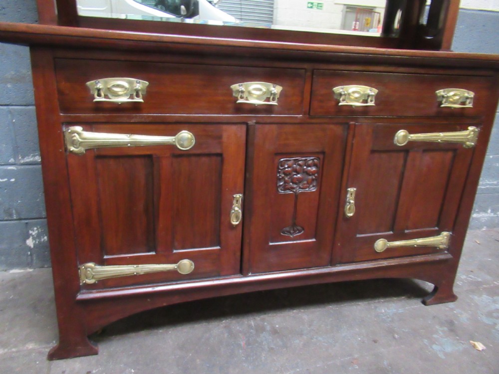 An Arts and Crafts period mahogany sideboard featuring leaded glass doors, brass strap hinges, a central carved tree motif, and a mirror back. View X