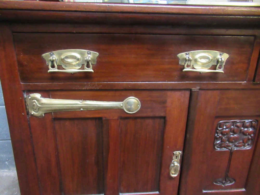 An Arts and Crafts period mahogany sideboard featuring leaded glass doors, brass strap hinges, a central carved tree motif, and a mirror back. View XI