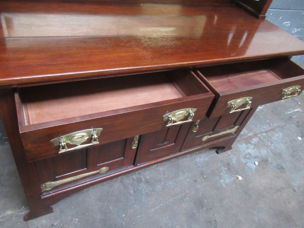An Arts and Crafts period mahogany sideboard featuring leaded glass doors, brass strap hinges, a central carved tree motif, and a mirror back. View XIV