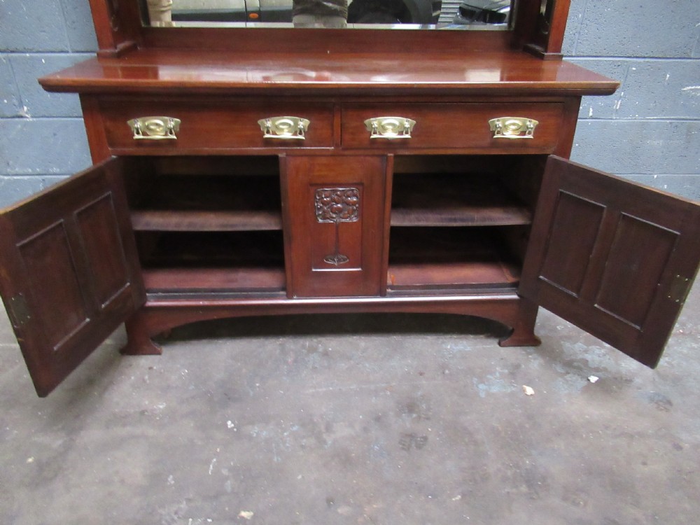 An Arts and Crafts period mahogany sideboard featuring leaded glass doors, brass strap hinges, a central carved tree motif, and a mirror back. View XV