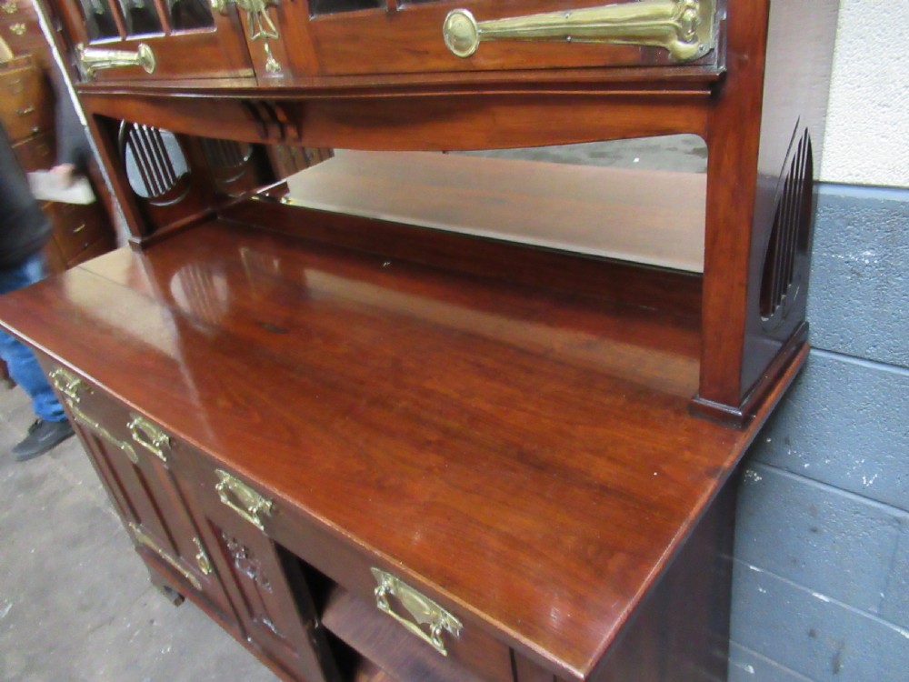 An Arts and Crafts period mahogany sideboard featuring leaded glass doors, brass strap hinges, a central carved tree motif, and a mirror back. View XVI