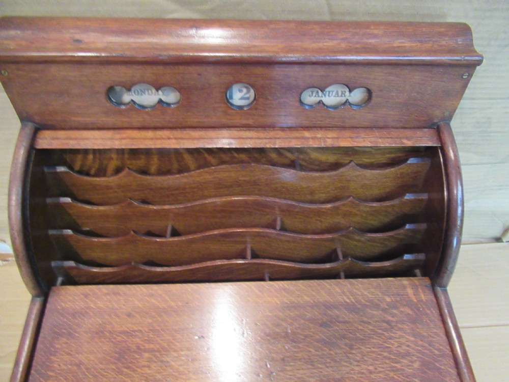 A late Victorian oak stationery cabinet featuring a tambour roll shutter, integrated perpetual calendar at the top, and a lower drawer with brass handle. View X