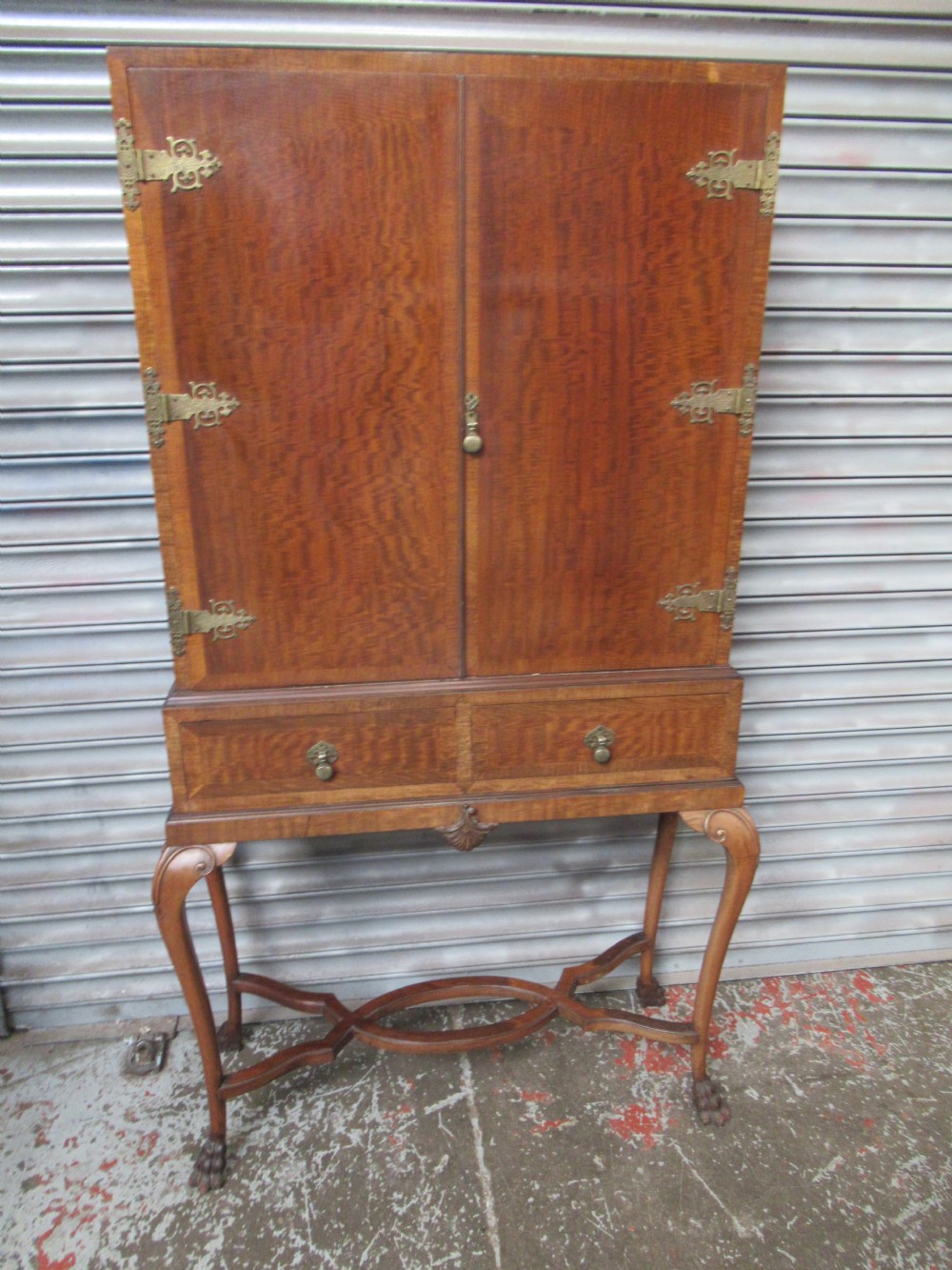 A 1930s mahogany cocktail cabinet on a stand featuring two upper doors with ornate brass strap hinges, lower drawers, cabriole legs, and claw and ball feet.