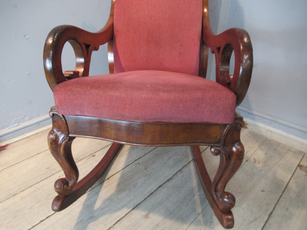 A Victorian mahogany rocking chair featuring carved scroll arms and dusty pink upholstery on curved wooden runners. View VI