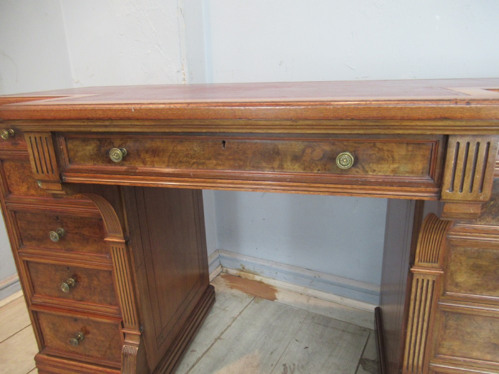 A late Victorian walnut pedestal desk featuring a dark leather writing inset, multiple burr walnut drawer fronts with brass knobs, and fluted column accents. View VIII