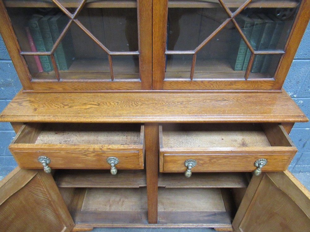 A 1920s warm brown oak cupboard bookcase featuring two upper glazed doors with geometric astragal glazing, two drawers, and a two-door lower cupboard. View XII