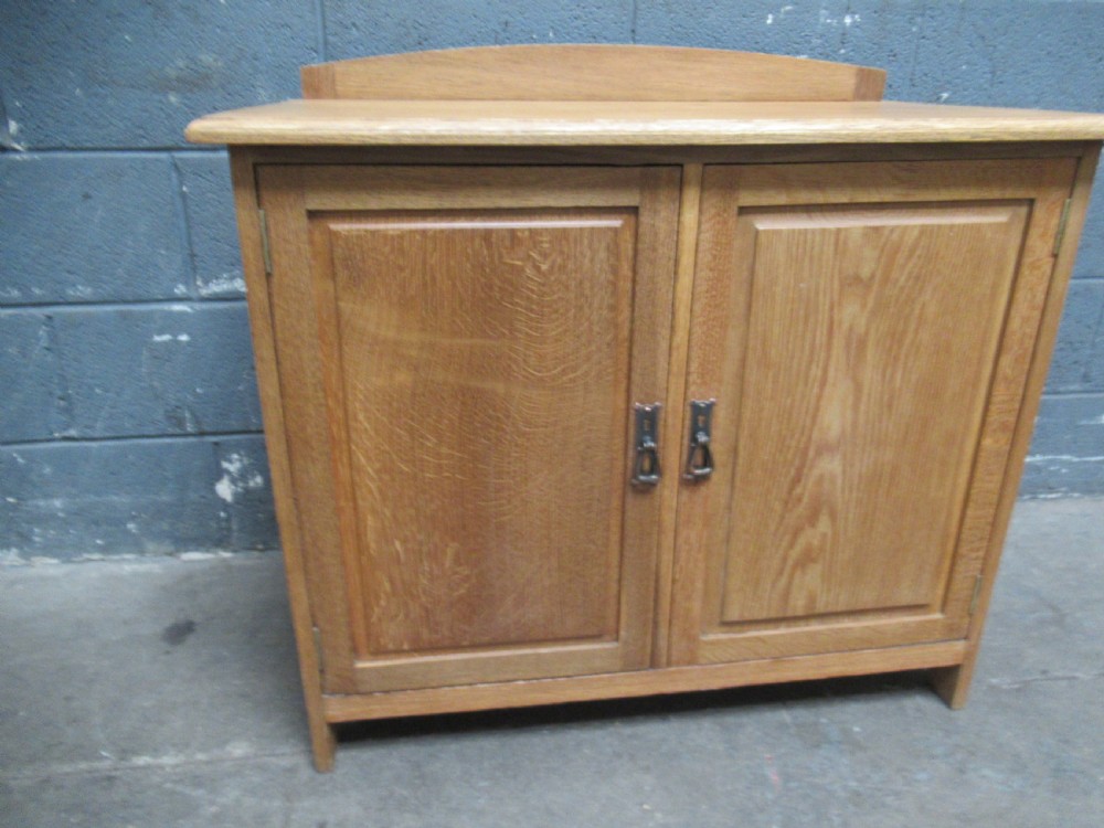 A light oak two-door storage cupboard featuring panelled doors, drop handles, and a low curved upstand along the top rear edge.