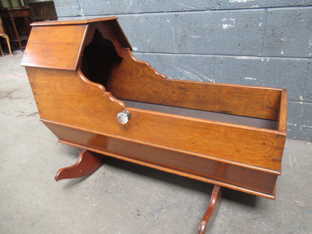 An antique Victorian oak rocking cradle with a hooded top, scalloped side rails, glass knob accent, and curved wooden rockers.