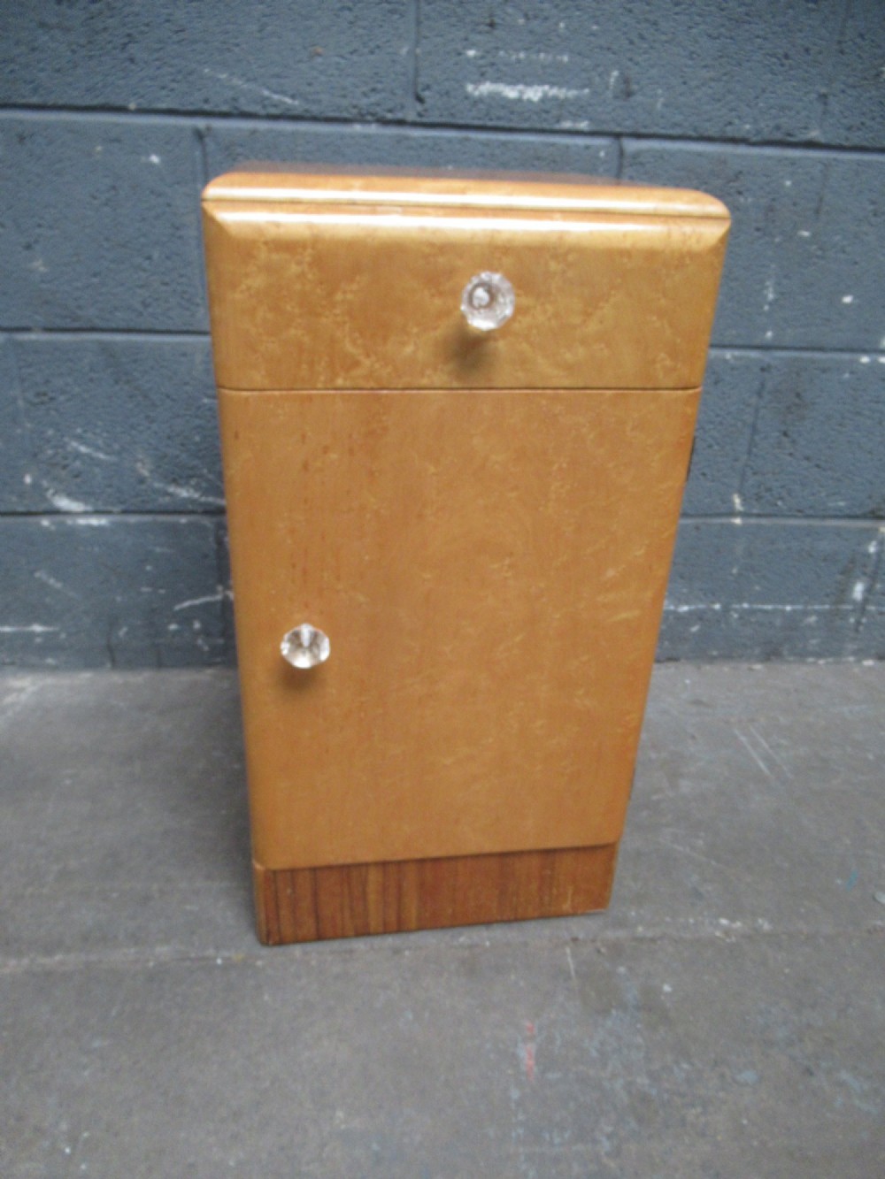 An Art Deco birds eye maple bedside cabinet with a rounded top, faceted glass knobs, single drawer, cupboard door, and darker wood plinth base.