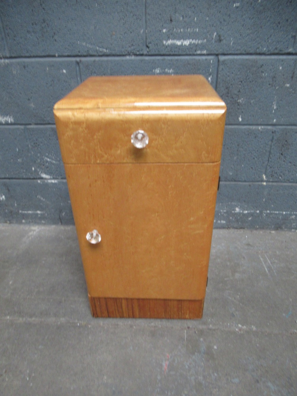 An Art Deco birds eye maple bedside cabinet with a rounded top, faceted glass knobs, single drawer, cupboard door, and darker wood plinth base. View II