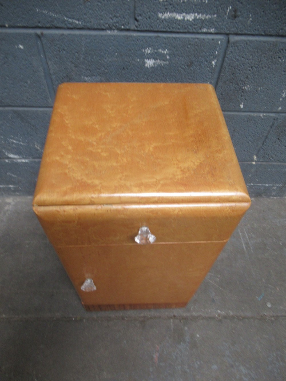 An Art Deco birds eye maple bedside cabinet with a rounded top, faceted glass knobs, single drawer, cupboard door, and darker wood plinth base. View IV