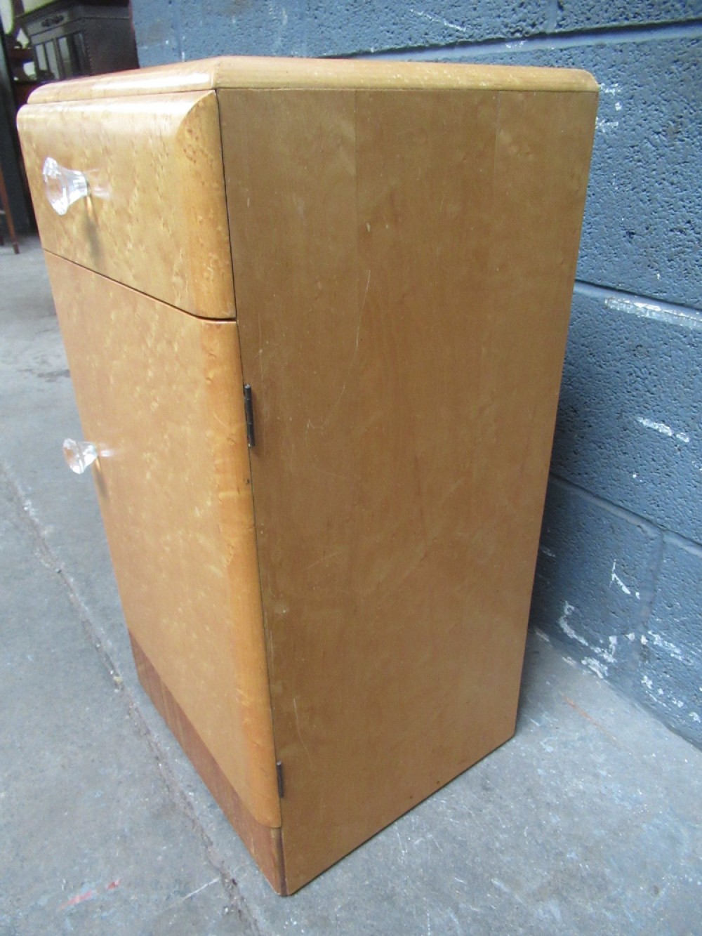 An Art Deco birds eye maple bedside cabinet with a rounded top, faceted glass knobs, single drawer, cupboard door, and darker wood plinth base. View V