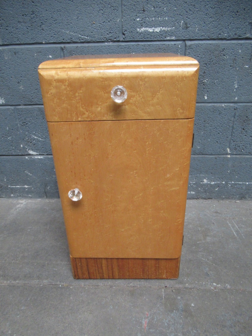 An Art Deco birds eye maple bedside cabinet with a rounded top, faceted glass knobs, single drawer, cupboard door, and darker wood plinth base. View IX