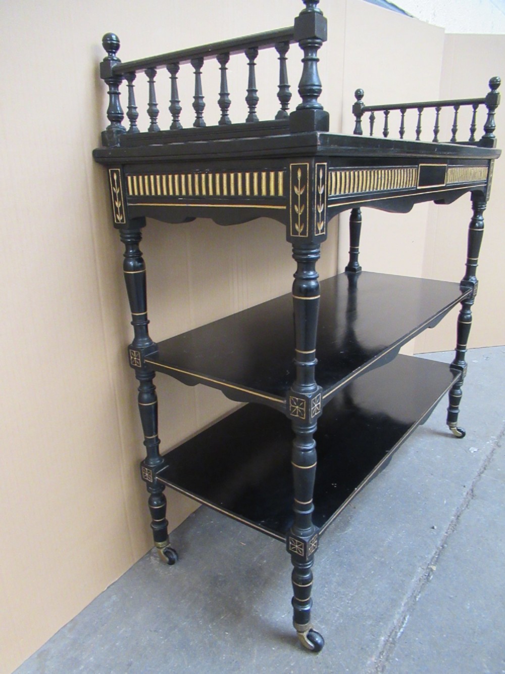 Victorian ebonised three-tier side table featuring spindle side galleries, turned legs, brass castors, and gold fluted detailing on the frieze. View IV