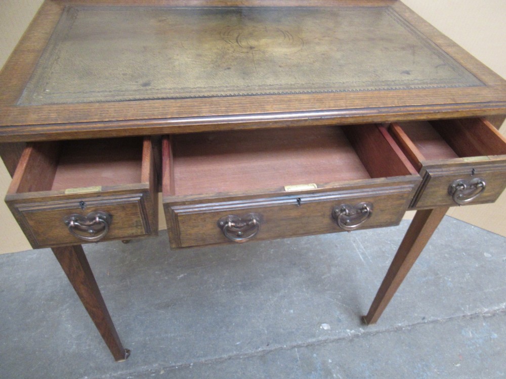 An Edwardian oak writing desk with a leather inset top, three drawers with brass handles, and a raised gallery back featuring a small bracket shelf. View X