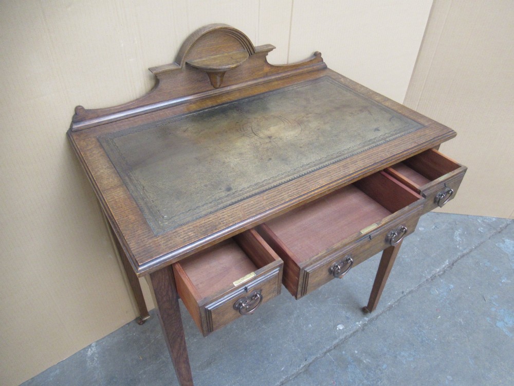 An Edwardian oak writing desk with a leather inset top, three drawers with brass handles, and a raised gallery back featuring a small bracket shelf. View XI