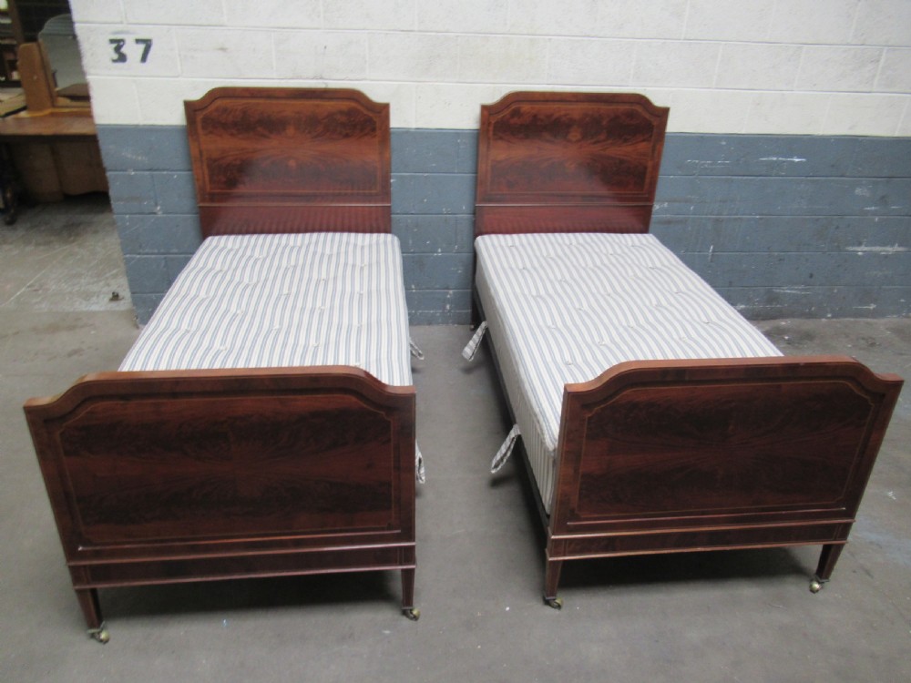 A pair of Edwardian mahogany single beds featuring flame mahogany panels, fine boxwood inlay, and tapered square legs with brass castors.