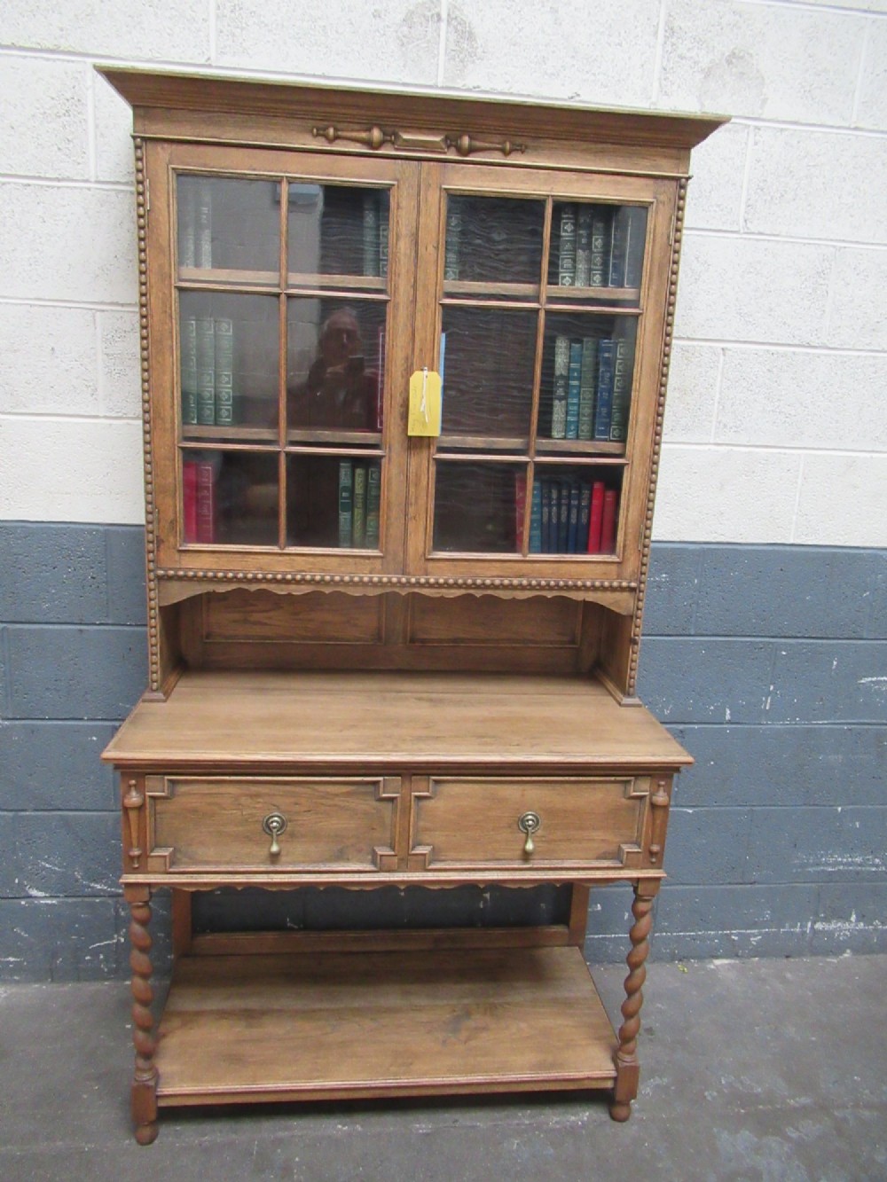 A 1920s oak two-door glazed bookcase featuring upper glass display cabinet, two geometric-moulded drawers, and barley twist legs with a lower undertier shelf.
