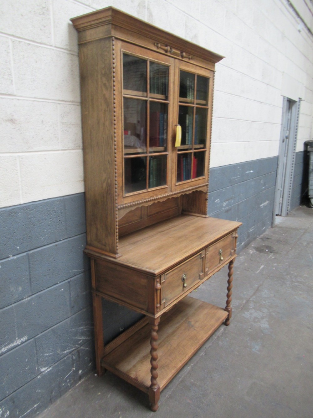 A 1920s oak two-door glazed bookcase featuring upper glass display cabinet, two geometric-moulded drawers, and barley twist legs with a lower undertier shelf. View II