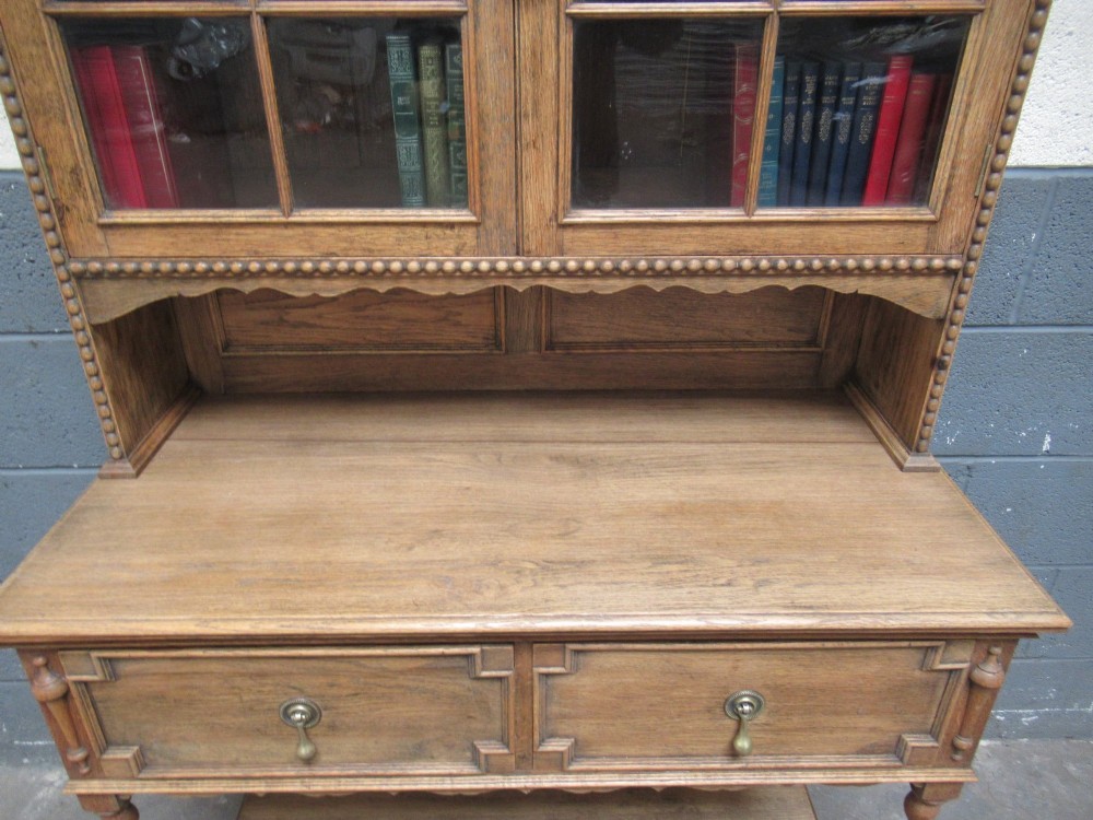 A 1920s oak two-door glazed bookcase featuring upper glass display cabinet, two geometric-moulded drawers, and barley twist legs with a lower undertier shelf. View VI