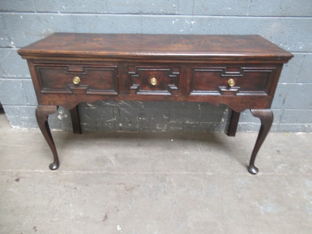 An antique dark oak dresser base featuring three drawers with geometric mouldings and brass knobs, supported by cabriole legs.
