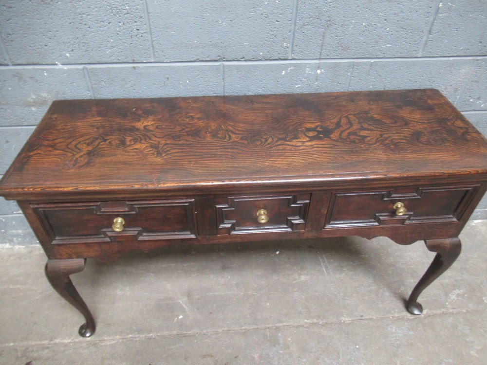 An antique dark oak dresser base featuring three drawers with geometric mouldings and brass knobs, supported by cabriole legs. View VII