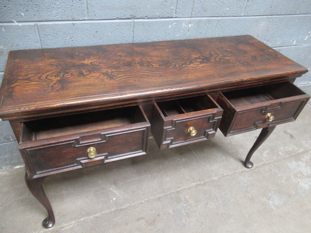 An antique dark oak dresser base featuring three drawers with geometric mouldings and brass knobs, supported by cabriole legs. View X