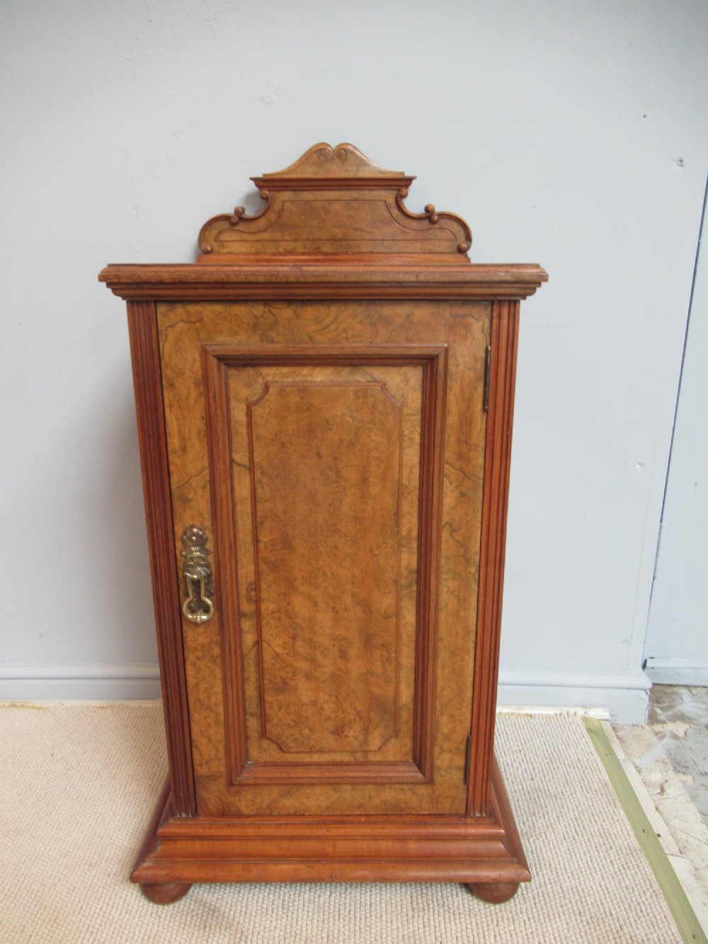 A late Victorian burr walnut bedside cabinet featuring a single panelled door with a brass drop handle, a shaped pediment gallery, and a plinth base on bun feet.