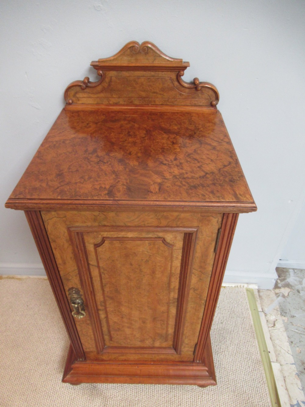 A late Victorian burr walnut bedside cabinet featuring a single panelled door with a brass drop handle, a shaped pediment gallery, and a plinth base on bun feet. View II